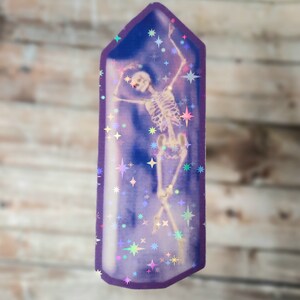 May include: A purple crystal-shaped sticker with a holographic finish features a skeleton dancing inside. The sticker is filled with iridescent stars.