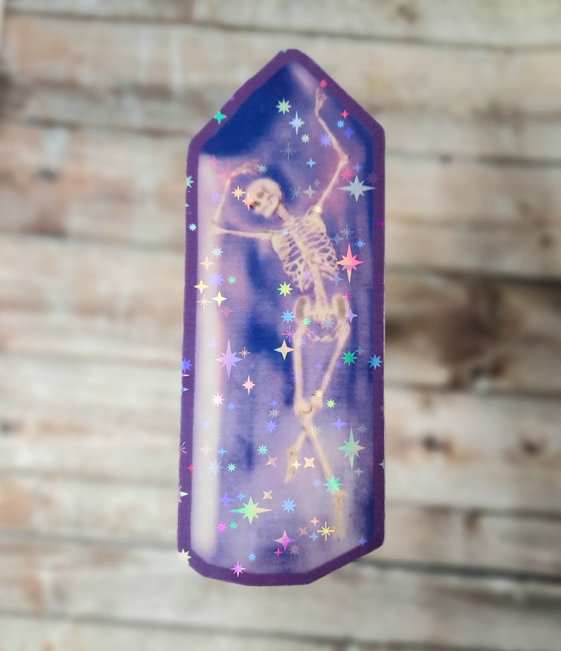 May include: A purple crystal-shaped sticker with a holographic finish features a skeleton dancing inside. The sticker is filled with iridescent stars.