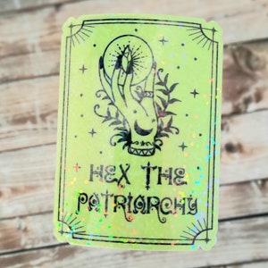 May include: A green holographic sticker with a black illustration of a hand holding a crystal ball. The text "HEX THE PATRIARCHY" is written below the illustration.