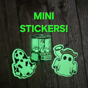 May include: Three glow-in-the-dark stickers featuring ghost designs. One sticker is a ghost wearing a cowboy hat, another is a ghost with polka dots, and the third is a ghost claw machine.