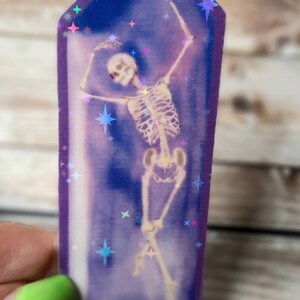 May include: A purple crystal-shaped sticker with a holographic finish features a skeleton dancing inside. The background is a dark blue with holographic stars.