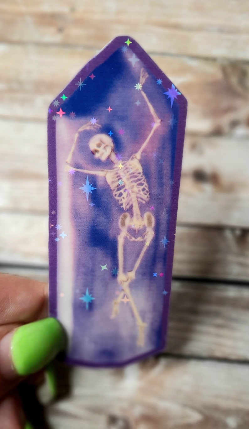 May include: A purple crystal-shaped sticker with a holographic finish features a skeleton dancing inside. The background is a dark blue with holographic stars.