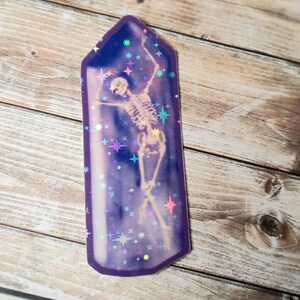 May include: A holographic sticker of a skeleton dancing inside a purple crystal with a glittery background.