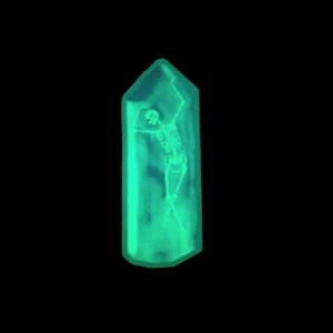May include: A green crystal-shaped pendant with a skeleton inside. The skeleton is in a dancing pose with its arms raised.