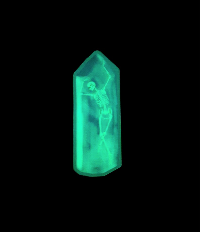 May include: A green crystal-shaped pendant with a skeleton inside. The skeleton is in a dancing pose with its arms raised.