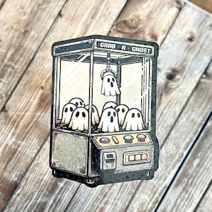 May include: A vintage-style claw machine with the text "Grab-A-Ghost" on the front. The machine is filled with white ghosts with black eyes and mouths.