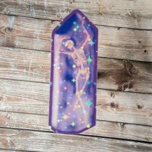 May include: A purple crystal-shaped sticker with a skeleton inside. The skeleton is dancing and there are iridescent glitter stars in the background.