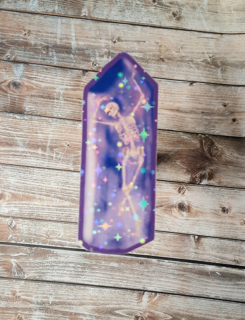 May include: A purple crystal-shaped sticker with a skeleton inside. The skeleton is dancing and there are iridescent glitter stars in the background.