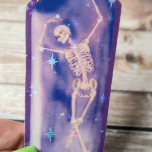 May include: A purple iridescent sticker featuring a skeleton dancing inside a crystal shape. The background is a dark blue with iridescent stars.