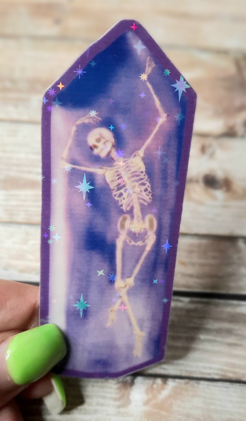 May include: A purple iridescent sticker featuring a skeleton dancing inside a crystal shape. The background is a dark blue with iridescent stars.
