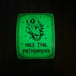 May include: A green glow-in-the-dark sticker with a black and white illustration of a hand holding a crystal ball. The text "HEX THE PATRIARCHY" is written below the illustration.