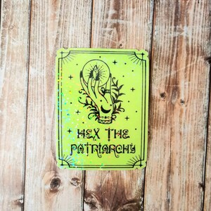 May include: A green holographic sticker with a black illustration of a hand holding a crystal orb. The text "HEX THE PATRIARCHY" is printed below the illustration.