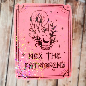 May include: Pink sticker with a black and white illustration of a hand holding a crystal ball. The text "HEX THE PATRIARCHY" is printed below the illustration.
