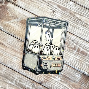May include: A black and white illustration of a claw machine with the text "Grab-A-Ghost" on the top. The claw machine is filled with white ghosts.