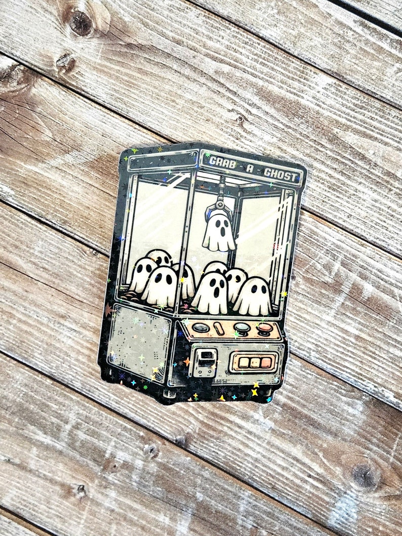 May include: A black and white illustration of a claw machine with the text "Grab-A-Ghost" on the top. The claw machine is filled with white ghosts.