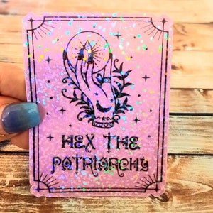 May include: Pink holographic sticker with a black outline and a hand holding a crystal ball. The text "Hex the Patriarchy" is printed in black.