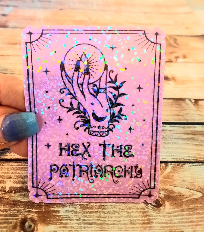 May include: Pink holographic sticker with a black outline and a hand holding a crystal ball. The text "Hex the Patriarchy" is printed in black.