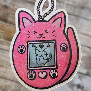 May include: Pink glitter cat-shaped keychain with a digital pet screen showing a cartoon cat. The screen has buttons with paw prints and a heart.