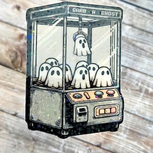 May include: A vintage-style claw machine with the text "Grab A Ghost" on the top. The machine is filled with white ghosts. The claw is holding a ghost.