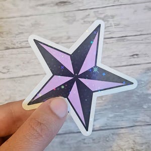 May include: A star-shaped sticker with a black and lavender design. The star has a white border and a holographic, glittery effect. The sticker is held by a hand.