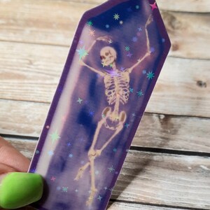 May include: A holographic sticker featuring a skeleton in a dancing pose inside a purple crystal shape with a glittery background.
