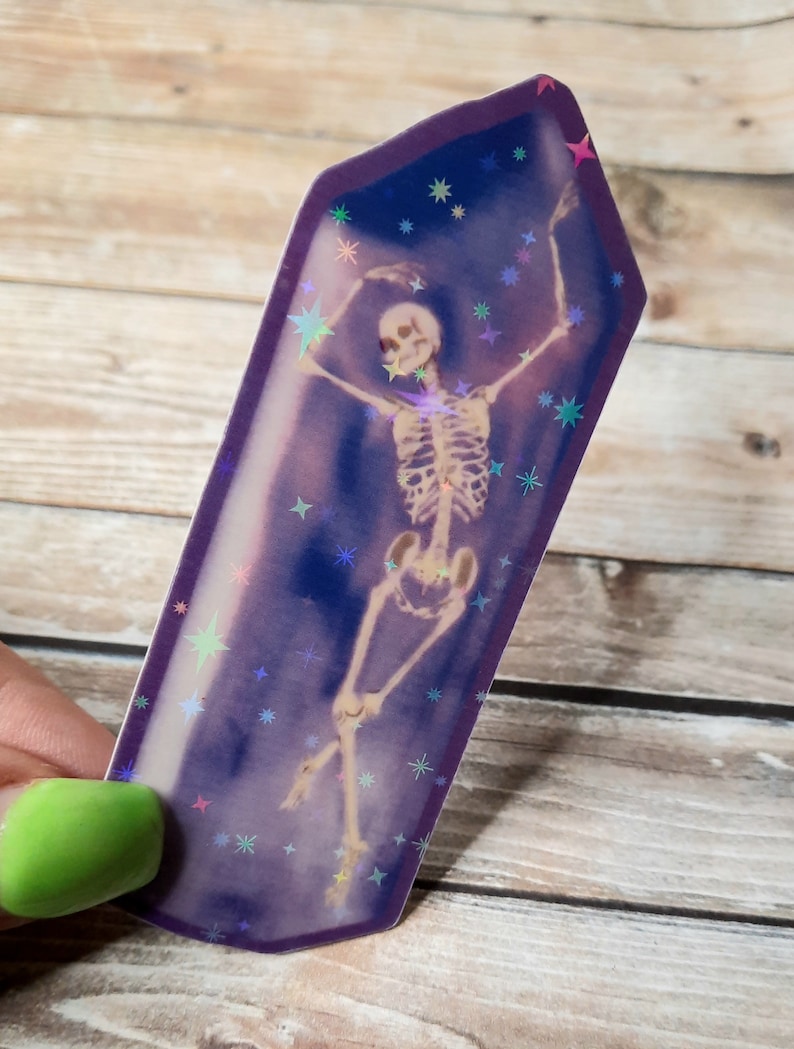May include: A holographic sticker featuring a skeleton in a dancing pose inside a purple crystal shape with a glittery background.