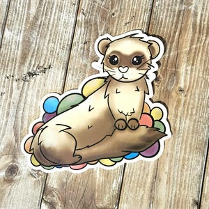 May include: A cartoon ferret with brown fur and big eyes sits on a bed of colorful rainbow-colored balls. The ferret has a pink nose and a white chest.