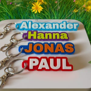 Könnte beinhalten: Vier farbige Namens-Schlüsselanhänger mit den Namen Alexander, Hanna, Jonas und Paul. Die Schlüsselanhänger liegen auf einer weißen Oberfläche mit einem Hintergrund aus grünem Gras und gelben Blumen.
