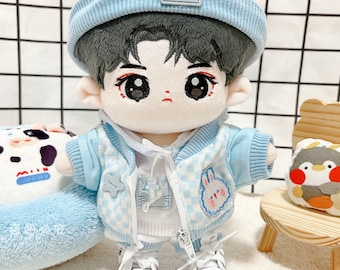 May include: A plush doll with a light blue and white checkered jacket, matching beanie, and white sneakers. The doll has dark hair, large eyes, and is wearing a white hoodie and pants. A bunny patch is on the jacket.