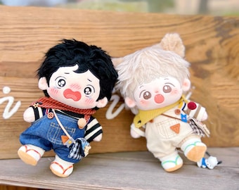 May include: Two plush dolls wearing overalls and scarves. The doll on the left has black hair and is wearing blue overalls. The doll on the right has blonde hair and is wearing beige overalls.