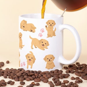 Könnte beinhalten: Eine weiße Kaffeetasse mit einem Muster aus Golden-Retriever-Welpen wird mit Kaffee aus einer Glaskanne gefüllt. Kaffeebohnen sind auf einer hellbeigen Oberfläche verstreut.
