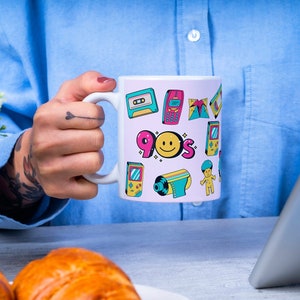 Pode incluir: Caneca de cerâmica branca com fundo rosa e ilustrações coloridas com tema dos anos 90, incluindo um rosto sorridente, uma fita cassete, um videogame e um telefone móvel. A caneca diz "90s" em letras rosa e amarelas.