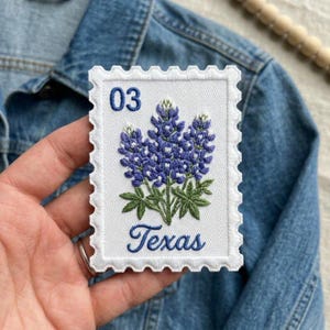 May include: A rectangular embroidered patch shaped like a postage stamp with a white background and scalloped edges. It features a blue lupine flower illustration, the number 03 in the top left corner, and the word Texas in blue script below.