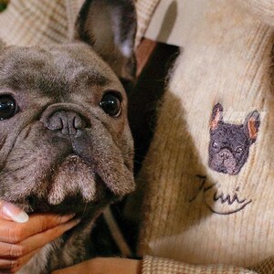 Puede incluir: Un bulldog francés gris es sostenido en brazos. El perro tiene ojos grandes y oscuros y una cara arrugada. La persona lleva un suéter beige con un pequeño diseño bordado de bulldog francés. El suéter tiene un patrón de cuadros.