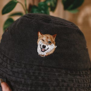 May include: A black bucket hat with a winking Shiba Inu dog embroidered on the front. The dog is depicted with brown and white fur, a black nose, and a collar. The hat is held by a hand, with green foliage in the background.