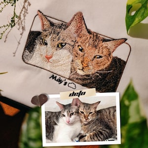 Puede incluir: Imagen bordada de dos gatos en una sudadera blanca. Los gatos son marrones y blancos con ojos negros. El texto "May's" está bordado debajo de los gatos. Una foto de dos gatos está pegada a la sudadera con el texto "defo" escrito en ella.