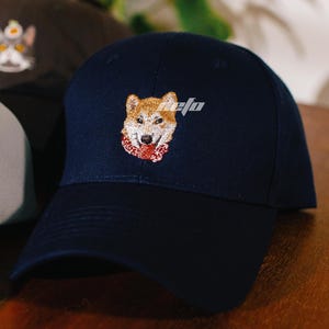 May include: A navy blue baseball cap with an embroidered image of a Shiba Inu dog wearing a red scarf.