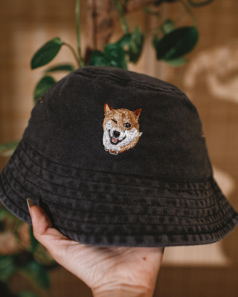 Puede incluir: Un sombrero de pescador negro con un Shiba Inu bordado en la parte delantera. El perro tiene pelaje marr&oacute;n y blanco, una nariz negra y un collar. El sombrero est&aacute; sostenido por una mano, con follaje verde en el fondo.