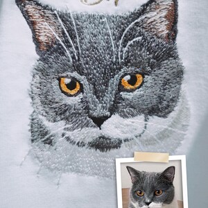 Puede incluir: Imagen bordada de un gato gris con ojos amarillos sobre un fondo blanco. El nombre del gato, "B.T.", est&aacute; bordado sobre la cabeza del gato en hilo dorado. Una foto del gato se muestra en un marco Polaroid debajo del bordado.