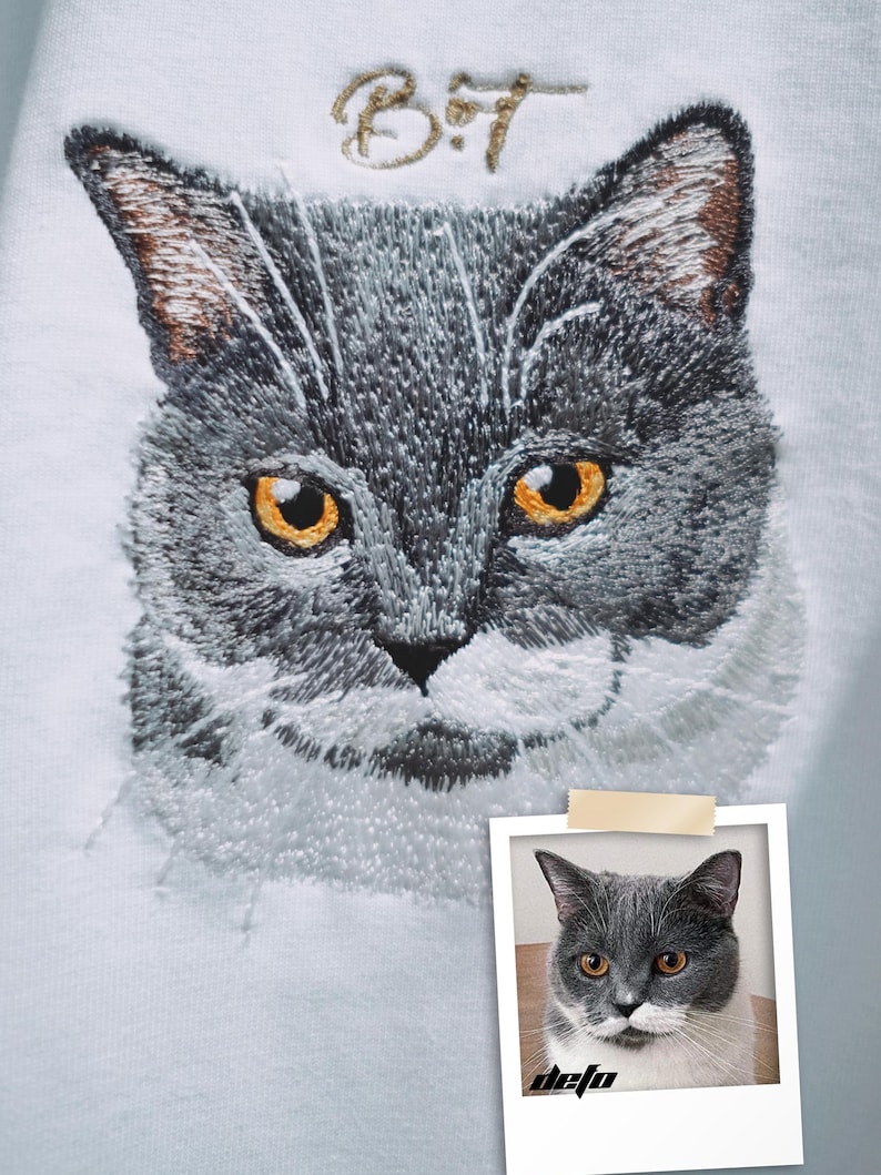 Puede incluir: Imagen bordada de un gato gris con ojos amarillos sobre un fondo blanco. El nombre del gato, "B.T.", est&aacute; bordado sobre la cabeza del gato en hilo dorado. Una foto del gato se muestra en un marco Polaroid debajo del bordado.