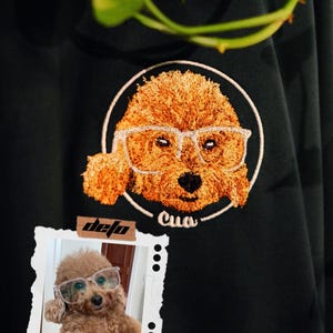 Puede incluir: Una camiseta negra con una imagen bordada de un perro con gafas. El perro es marr&oacute;n y tiene el nombre "Cua" bordado debajo. Una foto de un perro similar con gafas est&aacute; en la esquina inferior izquierda de la imagen.