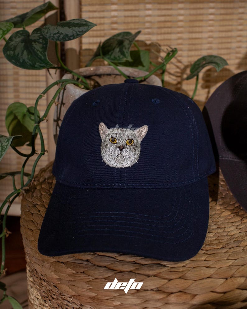 Puede incluir: Gorra de b&eacute;isbol azul marino con un bordado de la cara de un gato. La gorra tiene una visera curva y la palabra "defo" est&aacute; impresa en blanco. Un accesorio de moda.