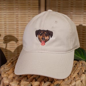 Puede incluir: Gorra de b&eacute;isbol beige con un rostro de perro bordado. El perro tiene pelaje marr&oacute;n y negro con una lengua roja. La gorra tiene la palabra "defo" bordada.