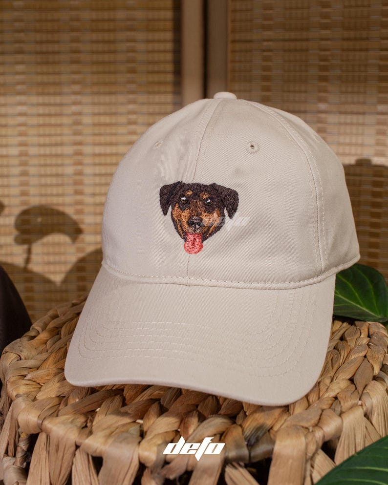 Puede incluir: Gorra de b&eacute;isbol beige con un rostro de perro bordado. El perro tiene pelaje marr&oacute;n y negro con una lengua roja. La gorra tiene la palabra "defo" bordada.