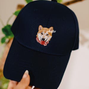 Puede incluir: Gorra de b&eacute;isbol azul marino con un Shiba Inu bordado con una bufanda roja. La gorra tiene una visera curva y una corona estructurada. La cara del perro est&aacute; detallada con hilo marr&oacute;n, blanco y negro, creando una apariencia realista.