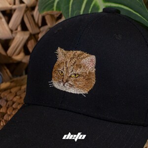 Puede incluir: Gorra de b&eacute;isbol negra con un rostro de gato bordado en tonos marrones y beige con ojos amarillos. La gorra tiene una visera curva y la palabra "defo" est&aacute; impresa en blanco en la parte delantera.