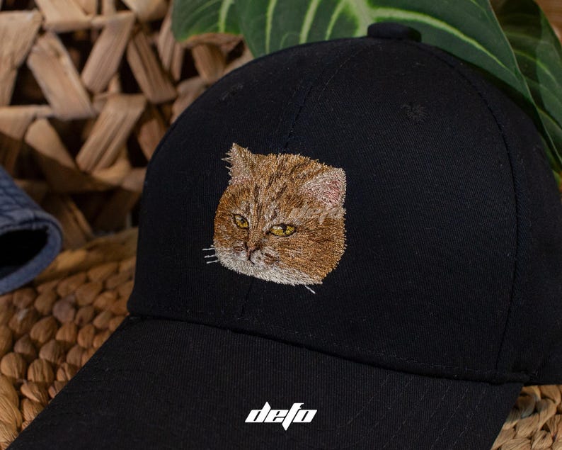 Puede incluir: Gorra de b&eacute;isbol negra con un rostro de gato bordado en tonos marrones y beige con ojos amarillos. La gorra tiene una visera curva y la palabra "defo" est&aacute; impresa en blanco en la parte delantera.