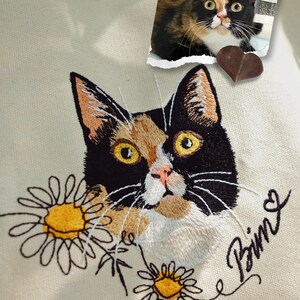 Puede incluir: Una bolsa de lona blanca con un retrato bordado de un gato negro y naranja. El gato est&aacute; rodeado de margaritas bordadas y el nombre "Bima" est&aacute; bordado en cursiva debajo del gato. Una peque&ntilde;a foto del gato est&aacute; unida a la bolsa con un coraz&oacute;n marr&oacute;n.