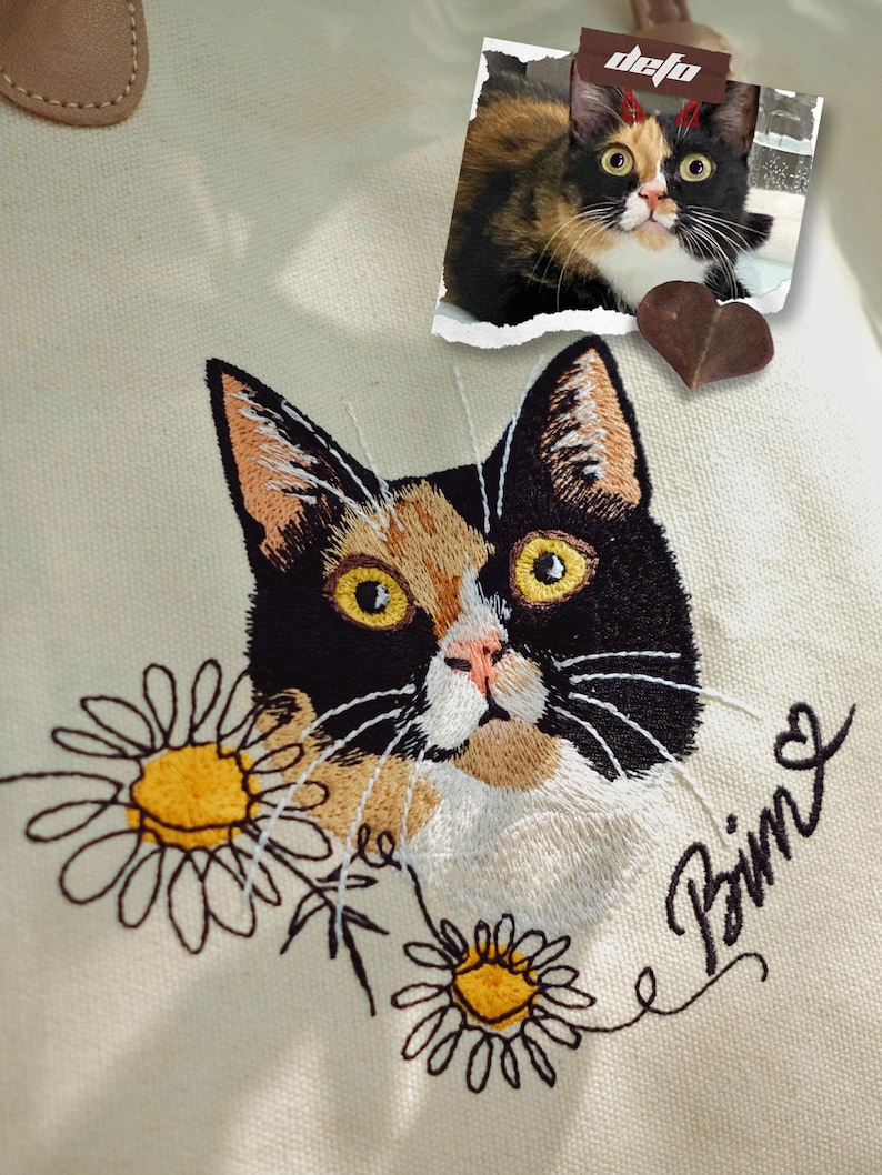 Puede incluir: Una bolsa de lona blanca con un retrato bordado de un gato negro y naranja. El gato est&aacute; rodeado de margaritas bordadas y el nombre "Bima" est&aacute; bordado en cursiva debajo del gato. Una peque&ntilde;a foto del gato est&aacute; unida a la bolsa con un coraz&oacute;n marr&oacute;n.