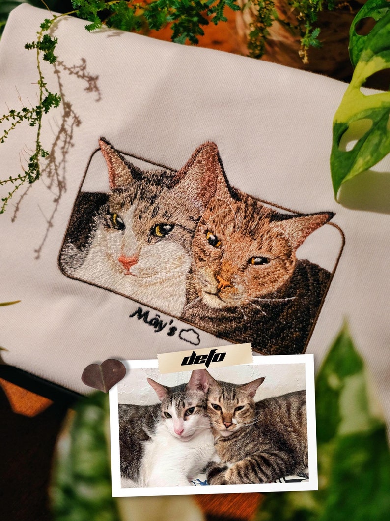 Puede incluir: Imagen bordada de dos gatos en una sudadera blanca. Los gatos son marrones y blancos con ojos negros. El texto "May's" est&aacute; bordado debajo de los gatos. Una foto de dos gatos est&aacute; pegada a la sudadera con el texto "defo" escrito en ella.